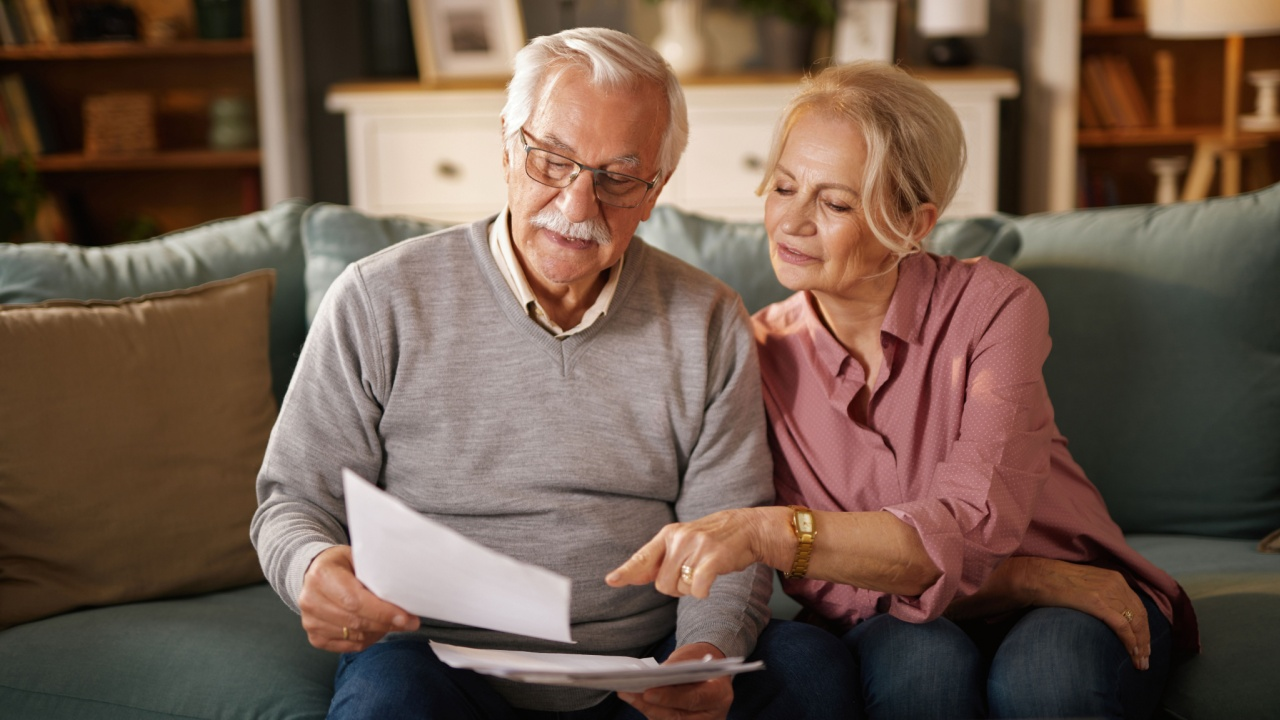 Older male/female couple looking at a piece of paper while sitting on a couch.