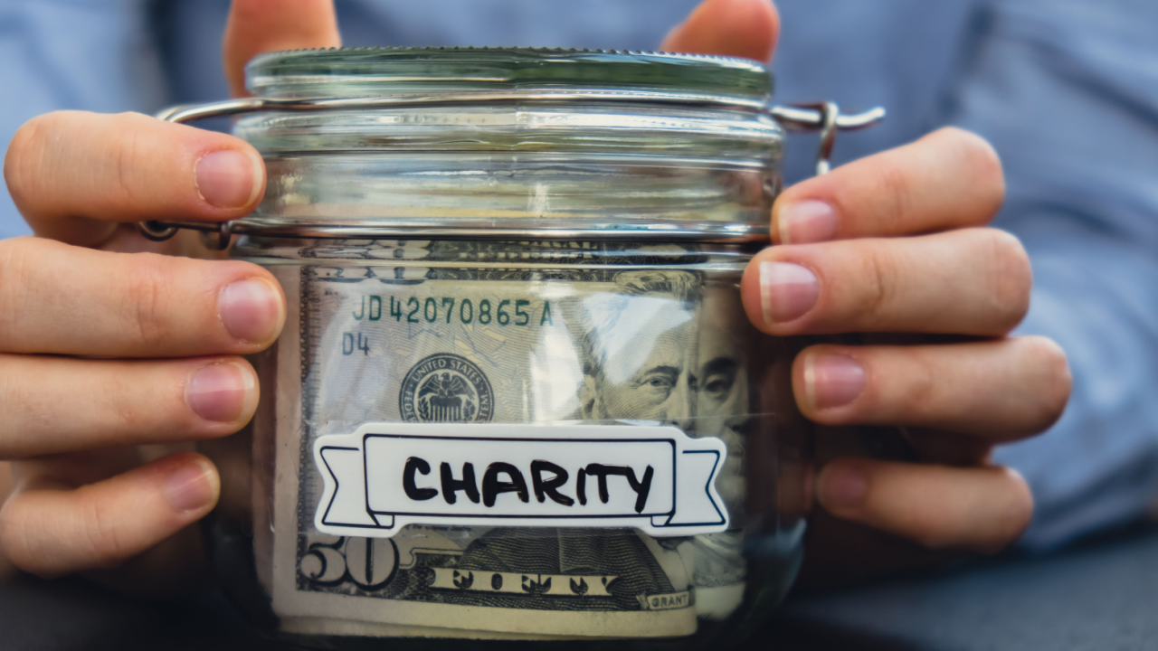 Two hands holding a glass jar with a label saying "Charity" on the jar. A US 50-dollar bill is visible inside the jar.